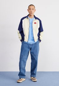 Mörkblå och elfenbensfärgad färgblockerad vindjacka med dragkedja och logotypmärke, i kombination med en ljusblå t-shirt, lösa denimjeans och bruna sneakers.