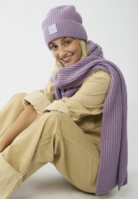 Lila gebreide beanie en sjaal, beide met een tekstuurpatroon, gecombineerd met een beige jumpsuit. Het model zit met gekruiste benen en lacht.