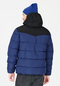 Blå och svart pufferjacka med huva, som har ett chevronmönster på axlarna och en quiltad textur. Mörka jeans bärs nedanför.