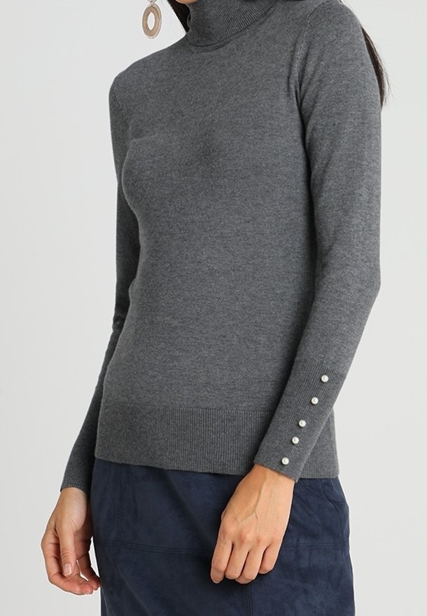 Femme portant un pull gris ajusté à col roulé avec des boutons en perle sur le poignet de la manche et une jupe bleu marine.