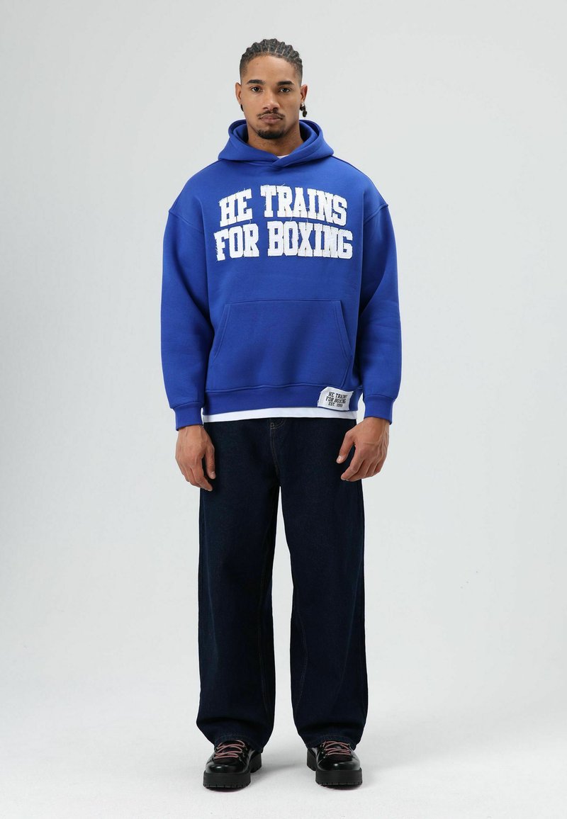 Koningsblauwe hoodie met witte tekst "HE TRAINS FOR BOXING," voorzak, gecombineerd met losse donkere spijkerbroek en zwarte schoenen.