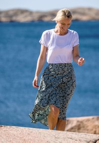 T-shirt blanc à manches courtes associé à une jupe à motifs dans des tons de vert et de gris, avec des motifs floraux et une fente sur le côté, au bord de la mer.