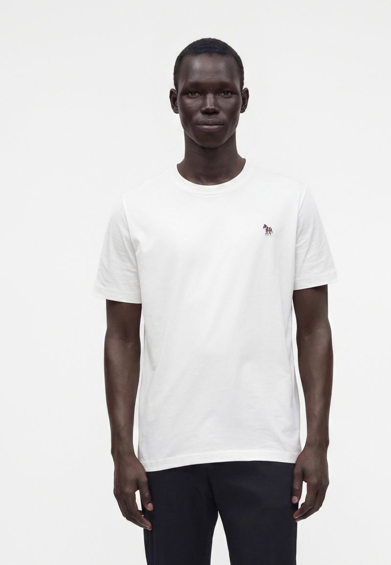 Jeune homme debout face à l'avant portant un t-shirt blanc uni à manches courtes avec un petit logo zèbre coloré sur la poitrine.