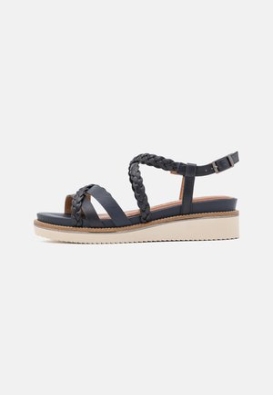 Sandale compensée noire avec brides tressées et plates, boucle à la cheville et semelle beige avec bande de roulement texturée pour une bonne adhérence.