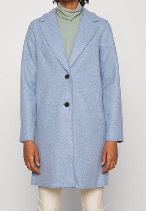 Hellblauer Wollmantel mit Revers und zwei schwarzen Knöpfen, getragen über einem grünen Rollkragenpullover und weißen Hosen.