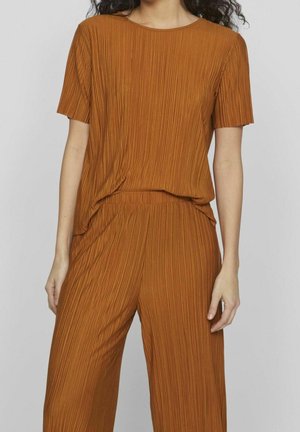 Femme portant un haut plissé à manches courtes couleur rouille et un pantalon ample plissé assorti sur un fond uni.