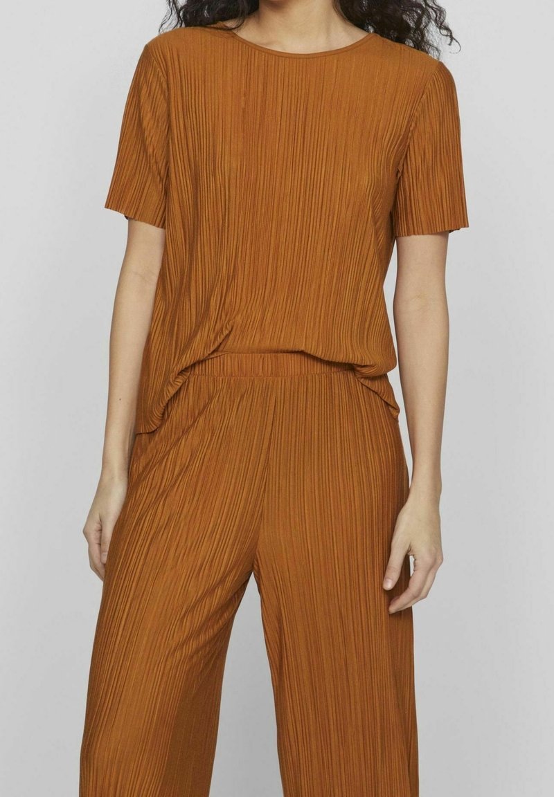 Femme portant un haut plissé à manches courtes couleur rouille et un pantalon ample plissé assorti sur un fond uni.