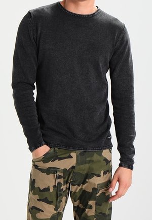 Mand iført en tætsiddende sort langærmet skjorte og camouflage cargobukser, med den ene hånd i lommen og den anden afslappet ved siden.