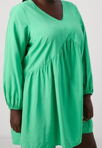 Grünes Langarmkleid aus glattem Stoff mit V-Ausschnitt, gerafftem Taillenbund und elastischen Ärmelbündchen, das eine lockere Passform und eine fließende Silhouette bietet.