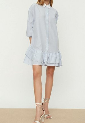 Femme portant une robe chemise à rayures bleu clair et blanc avec un ourlet volanté et des sandales blanches à talons hauts avec brides à la cheville.