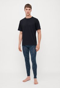 Schwarzes T-Shirt mit kurzen Ärmeln, kombiniert mit blauen gemusterten Leggings, die eine schlanke Passform und eine glatte Textur aufweisen, an einem männlichen Modell, das barfuß steht.