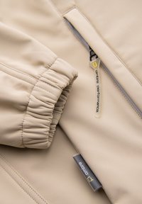 Manche de veste beige avec poignet élastique près d'une poche zippée avec une languette blanche étiquetée "ombre performance."
