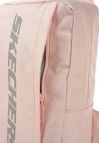 Mochila de tela rosa clara con un zipper lateral y un gran logo gris de "SKECHERS". Material texturizado y detalles de bolsillo adicionales visibles.