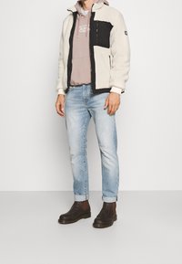 Giacca in pile color crema con dettagli neri, chiusura con zip e tasche laterali, indossata sopra un hoodie grigio chiaro e abbinata a jeans chiari e stivali scuri.