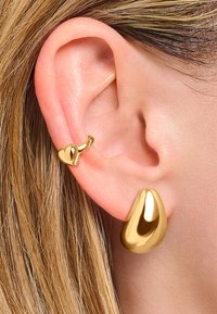 Goldene Tropfenohrringe mit einer glatten, reflektierenden Oberfläche, kombiniert mit einem kleinen, herzförmigen Stecker. Nahaufnahme in der Nähe eines Ohres.