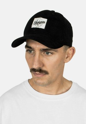 Cappello in corduroy nero con visiera arrotondata, caratterizzato da un'etichetta bianca con la scritta "Blackskies" in testo nero. Tessuto testurizzato con linee sottili.