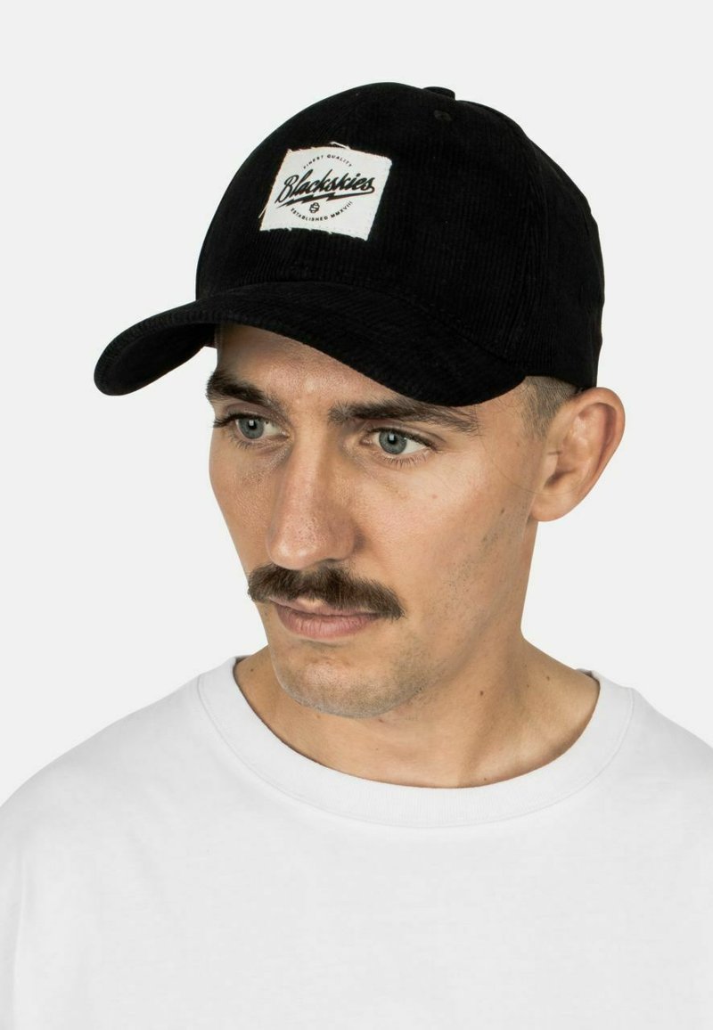 Zwarte corduroy cap met een ronde klep, voorzien van een witte patch met de tekst "Blackskies" in zwarte letters. Getextureerde stof met subtiele lijnen.