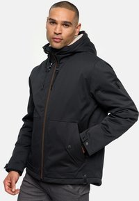 Chaqueta negra aislante con cremallera frontal, capucha ajustable, dos bolsillos laterales y un forro interior texturizado. Presenta detalles en cremallera marrón.