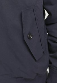 Navy blue jacket with a large patch pocket featuring a black button, smooth fabric, and elastic cuffs at the hem and sleeves.