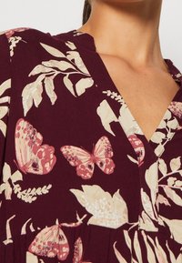 Burgundy blommigt klänning med ljusa blad och fjärilar. V-ringad halslinje och rynkad textur vid midjan.