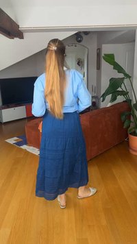 Donna con capelli lunghi biondi, camicetta blu e gonna blu scuro, in piedi a piedi nudi in un soggiorno con divano arancione e grande pianta verde.