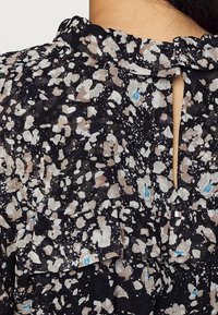 Gros plan sur un chemisier noir avec un motif floral abstrait beige, blanc et bleu, col à volants et détail dos avec ouverture en forme de goutte.