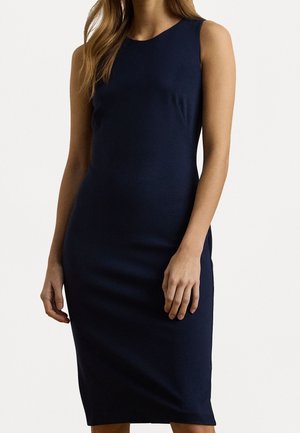 Mujer con un vestido azul marino sin mangas, ajustado y hasta la rodilla, de pie con los brazos relajados a los lados, sobre un fondo liso.