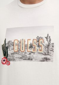 Camiseta blanca de algodón con un gráfico de paisaje y la palabra "GUESS" en bordado texturizado, junto con acentos florales en el lado izquierdo.