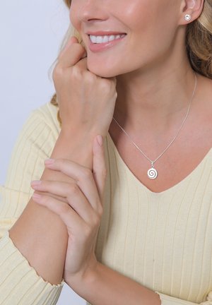 Silberne Spiral-Halskette an einer Frau, die ein hellgelbes, geripptes Oberteil trägt. Passende Spiral-Ohrringe in Form von Stecker. Glatte, polierte Oberfläche.