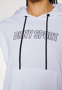 Gros plan sur une personne portant un sweat à capuche blanc avec des cordons noirs et l'inscription "DKNY SPORT" en lettres noires en gras sur la poitrine.
