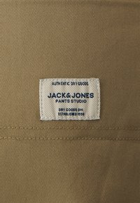 Beige stof med en vævet struktur har et syet rektangulært mærke med teksten "JACK & JONES PANTS STUDIO" i blåt.