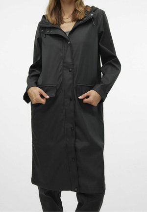 Femme portant un long imperméable noir avec capuche, boutons-pression à l’avant et deux poches à rabat, debout devant un fond uni.