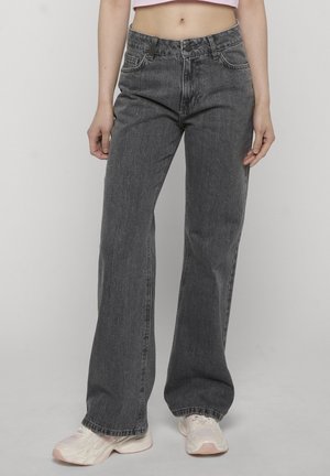 Persoon draagt vervaagde zwarte rechtgesneden jeans met hoge taille en witte sneakers, staand met ontspannen armen langs het lichaam tegen een effen achtergrond.