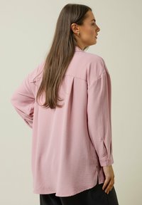 Camisa de manga larga rosa claro con un corte relajado, tejido texturizado, aberturas laterales y puños con botones, vista desde la parte de atrás.