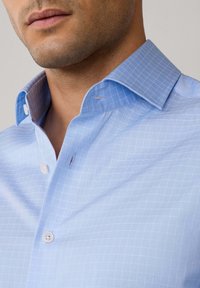 Camicia azzurra chiaro con un motivo a scacchi, colletto di un tono più scuro e bottoni bianchi. Tessuto di una trama liscia.