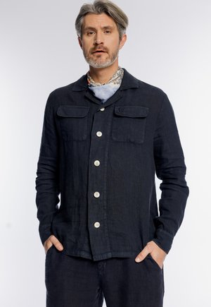 Mężczyzna z siwymi włosami i brodą, ubrany w ciemną zapinaną na guziki kurtkę z przednimi kieszeniami oraz wzorzysty szalik na szyi, z rękami w kieszeniach.