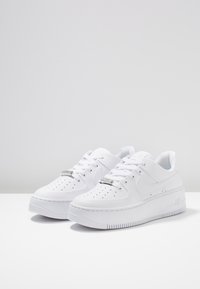 Para białych sneakersów Nike Air Force 1 typu low-top z perforowanym noskiem, białymi sznurowadłami i teksturowanymi gumowymi podeszwami na białej powierzchni.