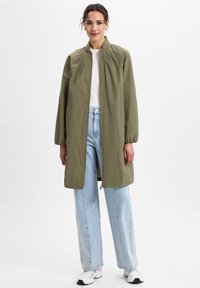 Olijfgroen lichtgewicht jack met een staande kraag, lange mouwen en een rits aan de voorkant, gecombineerd met lichtblauwe wijde jeans en witte sneakers.