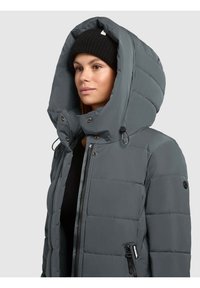 Veste matelassée grise avec une grande capuche surdimensionnée. Comprend des fermetures à boutons et à glissière, un bonnet en tricot noir à côtes, et une texture matelassée.