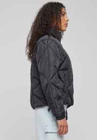 Juodas siūbuojantis pūkinis švarkas su aukštu apykakle, trumpu dizainu ir elastiniais rankogaliais. Yra priekis kišenė ir sklandus, blizgus audinio tekstūra.