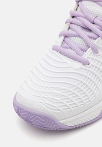 Sapata atlética branca com parte superior texturizada com detalhes em roxo, painéis de malha e sola com padrão em ziguezague para melhor aderência.