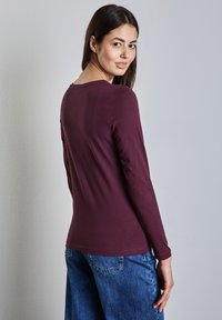 Bordeauxfarbene Langarmoberteil mit Rundhalsausschnitt, das eine glatte Textur aufweist. Kombiniert mit blauen Jeans im geraden Schnitt und mit Taschen.