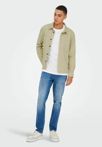 Hellbeige Hemd mit Knopfverschluss über einem weißen T-Shirt, kombiniert mit blauen Jeans und weißen Sneakers. Das Hemd hat eine Brusttasche und eine lässige Passform.