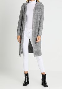 Długi szary dziergany kardigan z kapturem, ozdobiony teksturowanym wzorem i przednimi kieszeniami, noszony na białym golfie i białych dżinsach.