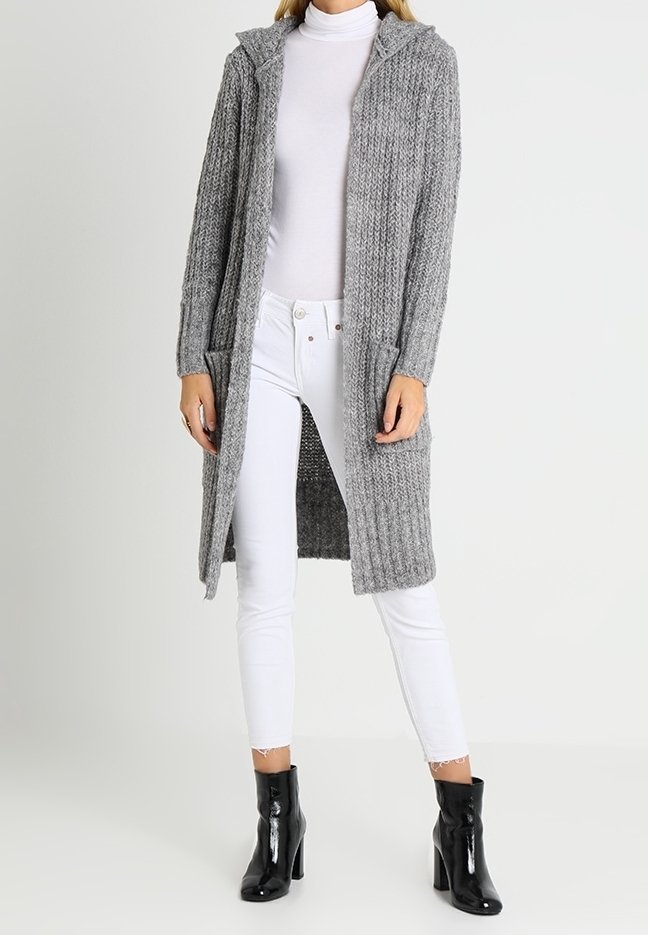 Długi szary dziergany kardigan z kapturem, ozdobiony teksturowanym wzorem i przednimi kieszeniami, noszony na białym golfie i białych dżinsach.