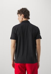 Schwarzes Polohemd mit Kragen, kurzen Ärmeln und glatter Textur. Verfügt über ein Logo auf dem linken Ärmel. Getragen mit roten Shorts.