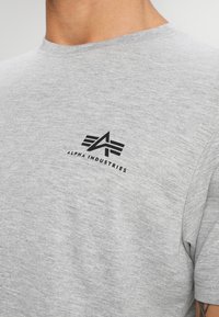 Camiseta de algodón gris de brezo con un pequeño logo negro de "Alpha Industries" en el área del pecho izquierdo. Diseño clásico de cuello redondo.