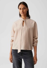 Light beige blouse made of textured fabric, featuring a high collar with a tie, long sleeves with elastic cuffs, and a keyhole detail.