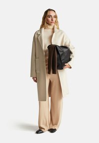 Cappotto in lana beige, dolcevita beige aderente, pantaloni a gamba larga e borsa in pelle nera con una sciarpa marrone drappeggiata all'interno. Texture morbide.