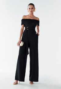 Sort off-shoulder jumpsuit med struktureret stof, vid ben-design, indsnøret talje med guldbælte, kombineret med en lille metallisk taske.